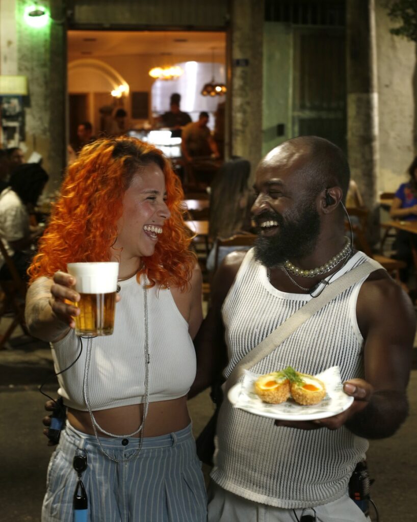 Uma mulher e um homem brindam em frente ao Bar Botica com o petisco em mãos